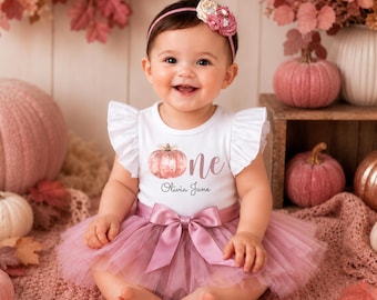 May include: A white short-sleeved bodysuit with ruffled shoulders, featuring a pink pumpkin graphic, the word one, and the name Olivia Jane. The bodysuit is paired with a dusty pink tulle skirt with a satin bow at the waist and a matching floral headband.