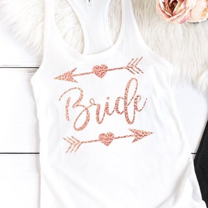 May include: White tank top with a rose gold glitter design that says "Bride" with arrows and hearts.