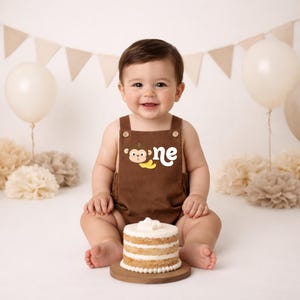 May include: A brown cotton romper featuring a graphic of a cartoon monkey face and two yellow bananas next to the white text "one" on the chest. The garment has two small wooden buttons at the top of the straps and a simple, solid-coloured design.