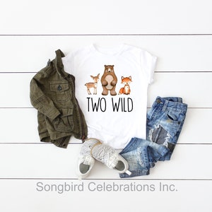 May include: White t-shirt with a brown bear, a deer, and a fox printed on it. The text "TWO WILD" is printed below the animals. The shirt is laid out on a white wooden surface with a green jacket, blue jeans, and white sneakers.