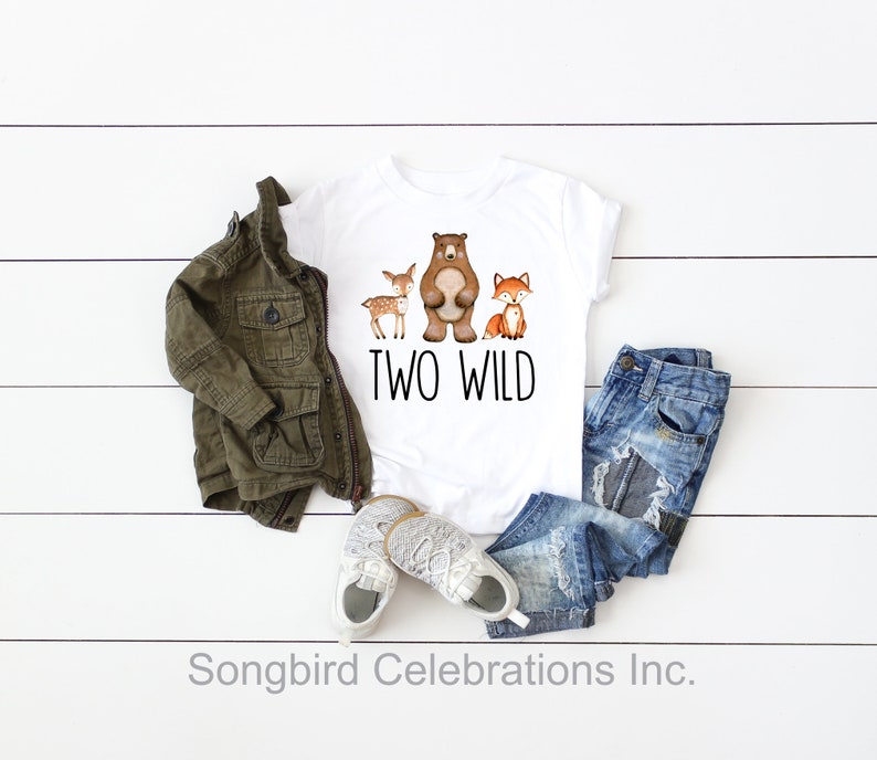 May include: White t-shirt with a brown bear, a deer, and a fox printed on it. The text "TWO WILD" is printed below the animals. The shirt is laid out on a white wooden surface with a green jacket, blue jeans, and white sneakers.