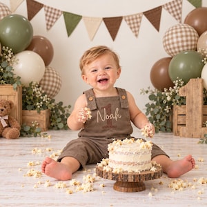 Puede incluir: Un ni&ntilde;o peque&ntilde;o con un peto marr&oacute;n con el nombre Noah y el n&uacute;mero 1 bordados, sentado ante un peque&ntilde;o pastel blanco sobre un soporte de madera. La escena incluye globos marrones y verdes, un oso de peluche y un n&uacute;mero 1 de madera.