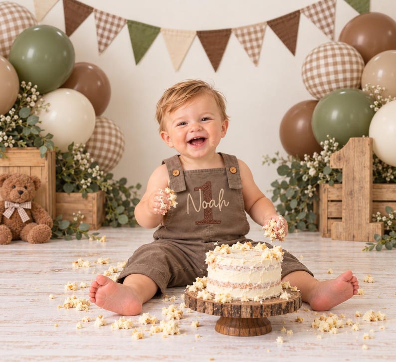 Puede incluir: Un ni&ntilde;o peque&ntilde;o con un peto marr&oacute;n con el nombre Noah y el n&uacute;mero 1 bordados, sentado ante un peque&ntilde;o pastel blanco sobre un soporte de madera. La escena incluye globos marrones y verdes, un oso de peluche y un n&uacute;mero 1 de madera.