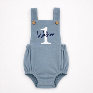 May include: A dusty blue corduroy romper featuring a large white number 1 and the name Walker embroidered in dark blue script. The garment has two shoulder straps with light brown wooden buttons and a snap closure at the bottom leg opening.