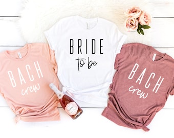 May include: Three t-shirts with text on them. One white t-shirt says "Bride to be" in black text. One peach t-shirt says "Bach crew" in white text. One maroon t-shirt says "Bach crew" in white text.