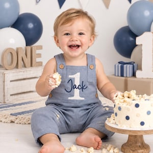 Puede incluir: Un peto de mezclilla azul claro con botones de madera, que presenta un n&uacute;mero uno bordado en blanco y el nombre Jack en letra cursiva azul oscuro. La prenda la lleva un ni&ntilde;o sentado junto a un pastel blanco con lunares azules.