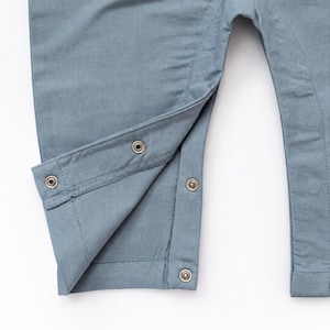 Puede incluir: Una prenda de color azul polvoriento que presenta una abertura en la pierna con cuatro broches met&aacute;licos. La tela tiene una textura tejida visible y el dise&ntilde;o incluye una costura reforzada a lo largo de la pierna interior.