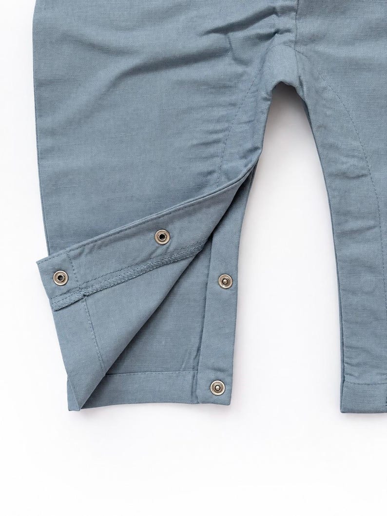 Puede incluir: Una prenda de color azul polvoriento que presenta una abertura en la pierna con cuatro broches met&aacute;licos. La tela tiene una textura tejida visible y el dise&ntilde;o incluye una costura reforzada a lo largo de la pierna interior.