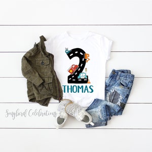May include: White t-shirt with a black number 2 on a road with cars and a helicopter. The name Thomas is written in teal below the number.