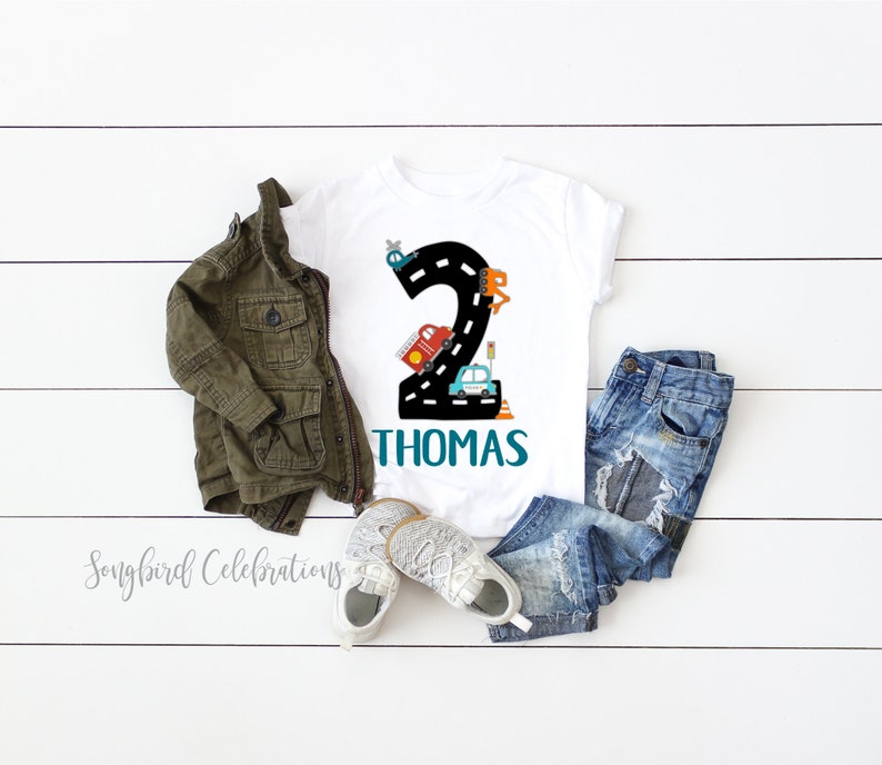 May include: White t-shirt with a black number 2 on a road with cars and a helicopter. The name Thomas is written in teal below the number.