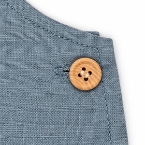 Puede incluir: Un primer plano de una prenda de tela azul con un bot&oacute;n redondo de madera clara con un patr&oacute;n de grano visible. El bot&oacute;n est&aacute; asegurado con hilo claro a trav&eacute;s de cuatro agujeros, colocado cerca de un ojal cosido en la tela.