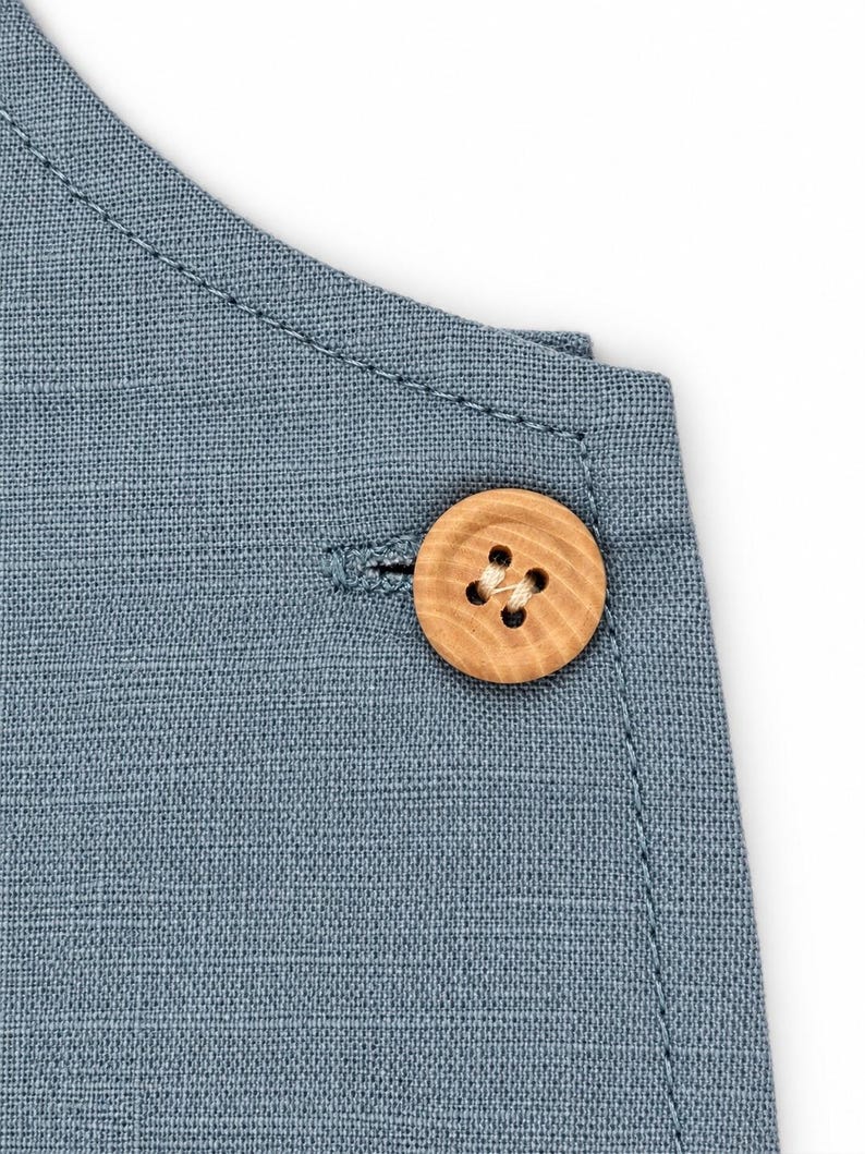Puede incluir: Un primer plano de una prenda de tela azul con un bot&oacute;n redondo de madera clara con un patr&oacute;n de grano visible. El bot&oacute;n est&aacute; asegurado con hilo claro a trav&eacute;s de cuatro agujeros, colocado cerca de un ojal cosido en la tela.