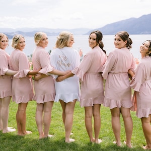 May include: Seven bridesmaids wearing pink robes with ruffled hems stand in a line, facing away from the camera. The bride is in the center wearing a white robe with the text "Mrs. Granier" in gold lettering.