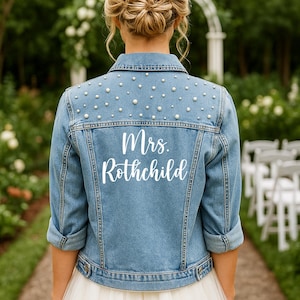 May include: A light blue denim jacket, adorned with pearls and the name "Mrs. Rothschild" in white script. The jacket, with rolled-up sleeves, is worn over a white tulle skirt, suitable for a wedding celebration.