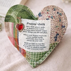 May include: A heart-shaped fabric prayer quilt featuring a patchwork design in green, beige, and multi-colored patterns. A white fabric patch is stitched to the center with black text and a small gold-toned charm with a red heart.
