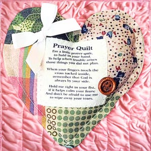 May include: A heart-shaped prayer quilt with a white bow and a button. The quilt is made of various patterned fabrics, including floral and polka dot designs. The text on the quilt reads "Prayer Quilt". The background is a quilted pink fabric.
