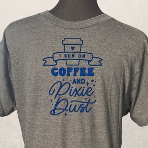 Op de afbeelding: Een grijs T-shirt met een blauwe afbeelding waarop staat "Ik draai op koffie en pixie dust". De afbeelding bevat een koffiebeker met een hartje erop.