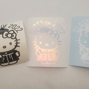May include: Three graduation stickers featuring Hello Kitty dressed in a graduation cap and gown holding a diploma. The stickers say "Class of 2022". The stickers are black, iridescent, and white.