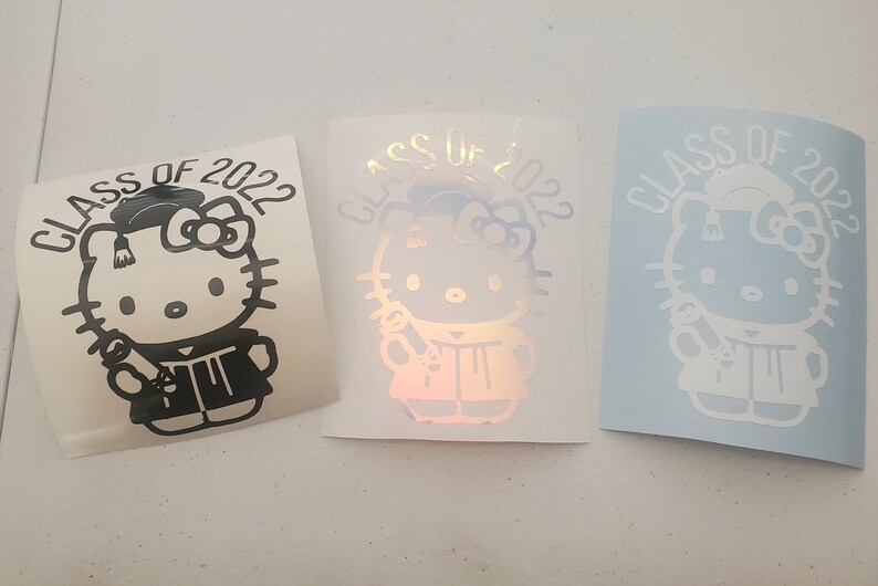 May include: Three graduation stickers featuring Hello Kitty dressed in a graduation cap and gown holding a diploma. The stickers say "Class of 2022". The stickers are black, iridescent, and white.