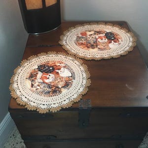 May include: Two round fabric doilies featuring a collage of various cat faces in shades of orange, brown, black, and white. Each doily has a cream-colored crocheted lace border with a scalloped edge and a secondary outer trim in light brown.