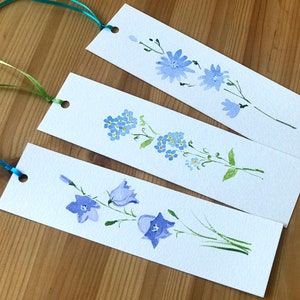 Könnte beinhalten: Drei rechteckige Lesezeichen aus weißem Papier, jedes mit einer individuellen Aquarell-Blumenillustration in Blau- und Grüntönen. Jedes Lesezeichen hat oben ein Loch mit einem dünnen Satinband in Türkis, Hellgrün oder leuchtendem Blau.