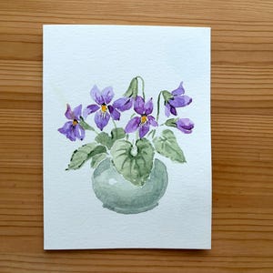 May include: Watercolour painting of a small vase with purple violets and green leaves. The vase is a soft green colour, and the flowers have yellow centres. The artwork is on a white card, set against a wooden background.