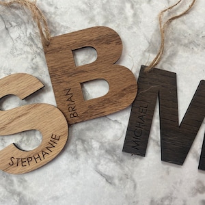 May include: Three wooden ornaments with the letters S, B, and M. The letters are painted in shades of brown and have the names Stephanie, Brian, and Michael engraved on them.