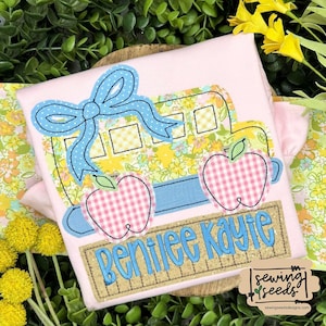 May include: A pink shirt with a yellow school bus, blue bow, and two red and white gingham apples. The bus is decorated with a floral pattern. Below the bus is a brown and gold sign with the name "Bentilee Kayle" in blue lettering. The shirt is for a child.
