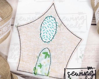 May include: A beige fabric church applique with a cross on top. The church has two windows, one with a teal and white pattern and the other with a green and white floral pattern. The fabric is stitched with black thread. The text "sewing seeds" is in black lettering on a white background.