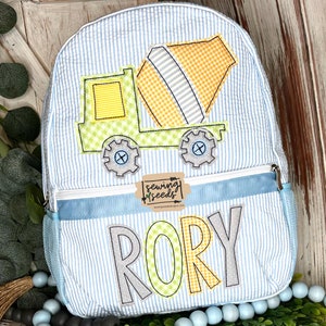 Peut inclure: Un sac à dos rayé bleu et blanc avec une applique de camion à béton jaune et vert. Le sac à dos a une poche zippée bleue et une poche latérale en filet. Le nom "RORY" est brodé sur le devant du sac à dos en lettres jaunes, vertes et grises.