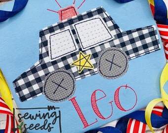 May include: A blue and white checkered police car applique with a yellow star and the name Leo embroidered in pink on a light blue fabric. The applique is surrounded by blue, yellow, and red ribbons.