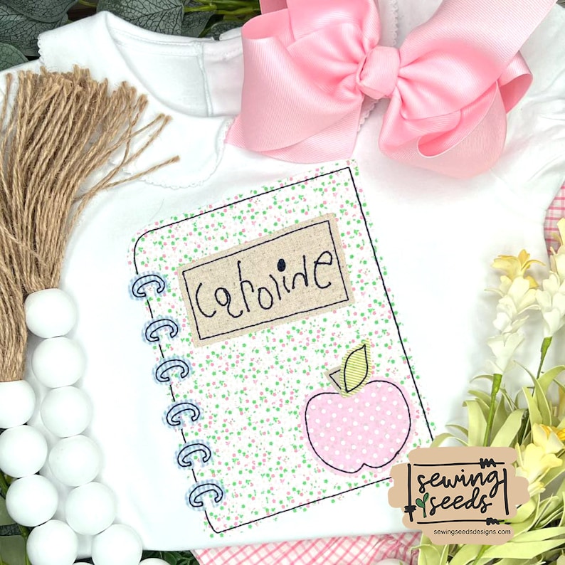 May include: A white onesie with a pink bow and a stitched design of a notebook with the word "Caroline" on it. The notebook has a pink apple with a green leaf on it. The onesie is surrounded by white beads and a brown rope.