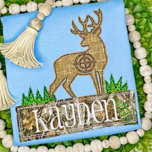 May include: A light blue t-shirt with a brown deer silhouette with a target on its back. The deer is standing in green grass. The t-shirt has a camouflage fabric patch with the name "Kayden" in white text.