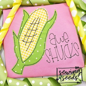 May include: A pink t-shirt with an embroidered corn on the cob and the text "Awe Shucks". The corn is yellow and green with a white and yellow checkered pattern. The text is in a handwritten style. The t-shirt is surrounded by green and yellow ribbon with white polka dots.
