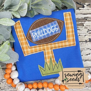 May include: A blue t-shirt with an orange and white checkered football goal post and a brown football with the word "BULLDOGS" on it. The football goal post is sitting on top of green grass. The t-shirt has a brown label with the text "sewing seeds" and the website address "sewingseedsdesigns.com".
