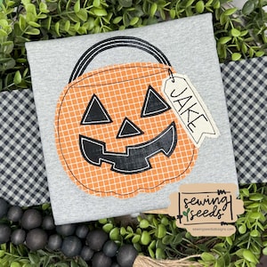 May include: A gray t-shirt featuring a large orange pumpkin design with a black handle and face. A tag with the name "JAKE" hangs from the pumpkin. The shirt has a Halloween theme.