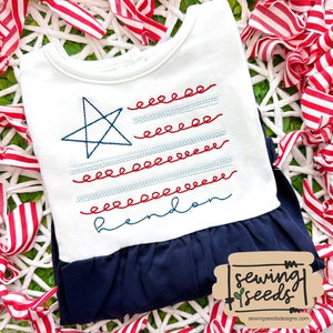 May include: White and navy blue baby outfit with an embroidered American flag design. The flag features a blue star and red and blue stitched stripes. The name "hendon" is embroidered in blue script. The outfit is from Sewing Seeds Designs.