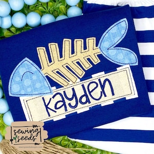 May include: A blue and white striped shirt with a blue fish bone applique and the name "Kayden" embroidered on a beige banner.