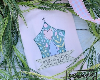 May include: A pink fabric with a green and blue embroidered house with a pink heart and a banner that says "Love lifted me". The fabric is surrounded by green foliage.