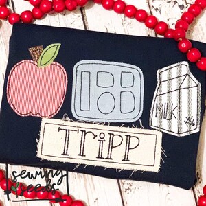 Könnte beinhalten: Ein marineblaues T-Shirt mit einer gestickten Apfel, Lunchbox und Milchpackung. Der Name "Tripp" ist auf einem beigefarbenen Rechteck gestickt.