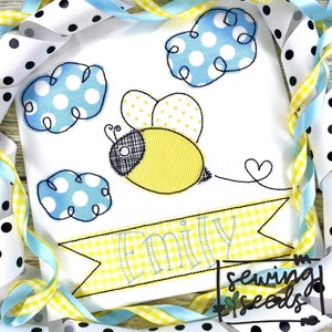 May include: A yellow and black embroidered bee with a striped body and polka dot wings flying above three blue clouds with white polka dots. A yellow banner with a gingham pattern and the name "Emily" is below the bee. The text "sewing seeds" is in the lower right corner of the image.
