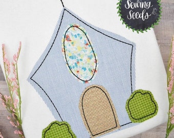 May include: Embroidered design of a church building with a cross on top, a stained glass window, a door, and two green bushes. The design is stitched on a white fabric.