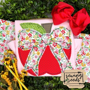 May include: A light pink toddler shirt with an apple design. The apple is red with a white polka dot pattern and a floral bow. A large red bow is placed on the top right. The shirt is surrounded by floral fabric and greenery.