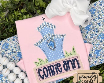 May include: A pink baby onesie with an embroidered blue and white church design and the name "Cobie Ann" on a tan banner. The onesie is surrounded by white beads and a white bow.