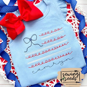 May include: A light blue sleeveless top with a red, white and blue embroidered design of an American flag with a bow on top. The top has a ruffled trim around the neckline and armholes. The text "Carolyn" is embroidered below the flag.
