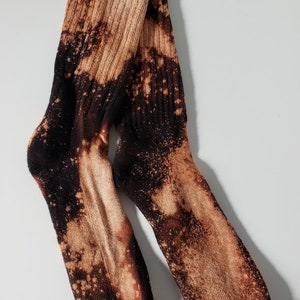 Może przedstawiać: Para czarno-brązowych skarpet z wzorem rozpryskanym wybielaczem. Skarpety są wykonane z miękkiego, wygodnego materiału i mają żebrowaną fakturę.