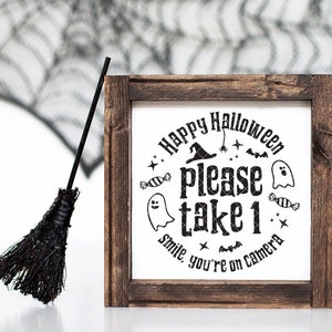 May include: A wooden sign with a white background and black text that reads "Happy Halloween Please Take 1 Smile, You're On Camera". The sign is decorated with a witch's hat, bats, candy, and ghosts.