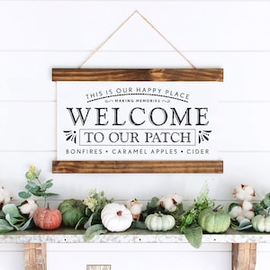 Könnte beinhalten: Ein Holzschild mit dem Text "This is our happy place making memories. Welcome to our patch. Bonfires, caramel apples, cider." Das Schild hängt an einer weißen Wand mit einem Regal darunter. Das Regal ist mit Herbstdekorationen geschmückt, darunter Kürbisse, Wattebäusche und Grünpflanzen. Das Schild ist von Woodworks hergestellt.