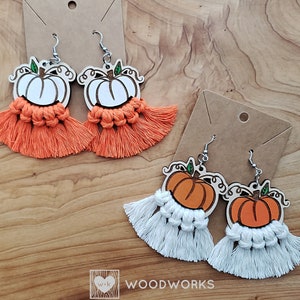 May include: Two pairs of pumpkin-shaped earrings with tassels. One pair has orange tassels and the other pair has white tassels. The earrings are made of wood and have a laser-cut design. The text "WOODWORKS" is visible in the image.