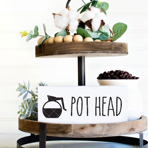May include: A two-tier wooden tiered tray with a black metal stand. The top tier has a white sign with a black coffee pot graphic and the text "POT HEAD". The bottom tier has a white bowl with coffee beans.